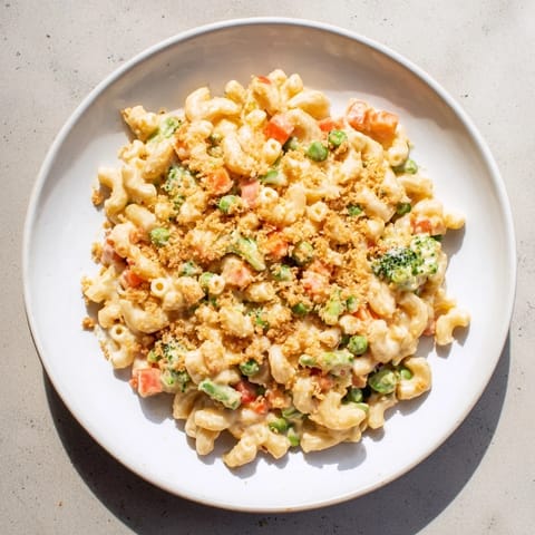 A close-up of baked Garden Veg Mac and Cheese bubbling in a dish, topped with golden panko crumbs and colorful peas, broccoli, and carrots.